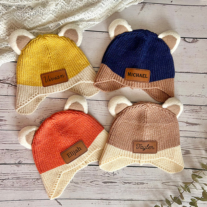 A collection of four personalized baby beanie hats with cute bear ears, available in yellow, blue, brown, and orange colors, each featuring a different name on the leather patch.