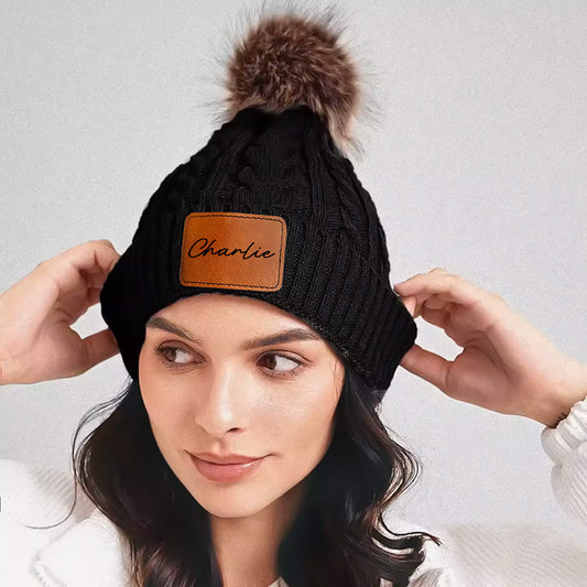 A woman wearing a black cable knit beanie with a brown faux fur pom and a brown custom leather patch with the name on it.
