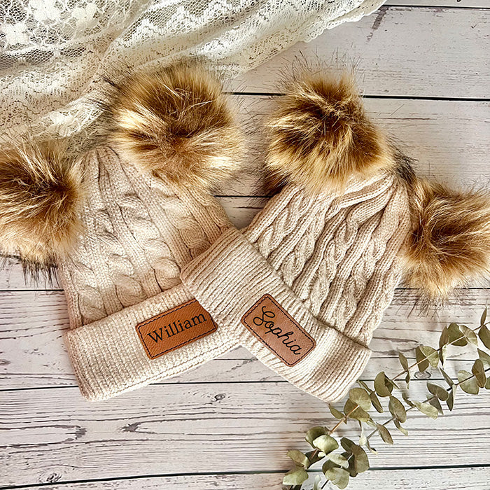 Children's beanie hats with double pom poms, beige color, both with a personalized leather patch on the front.