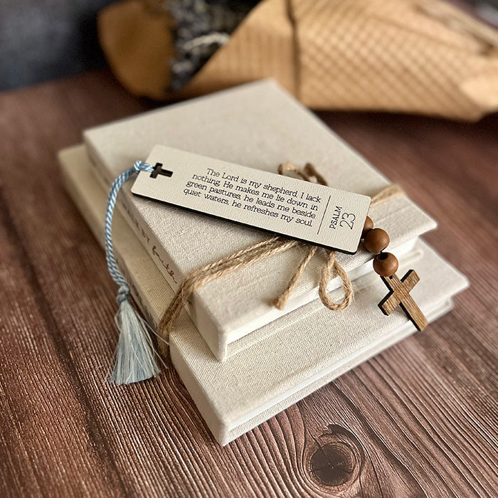 A collection of wooden bookmarks with white textured surfaces and Bible verses engraved on them, displayed on a wooden surface with a cross and a tassel.