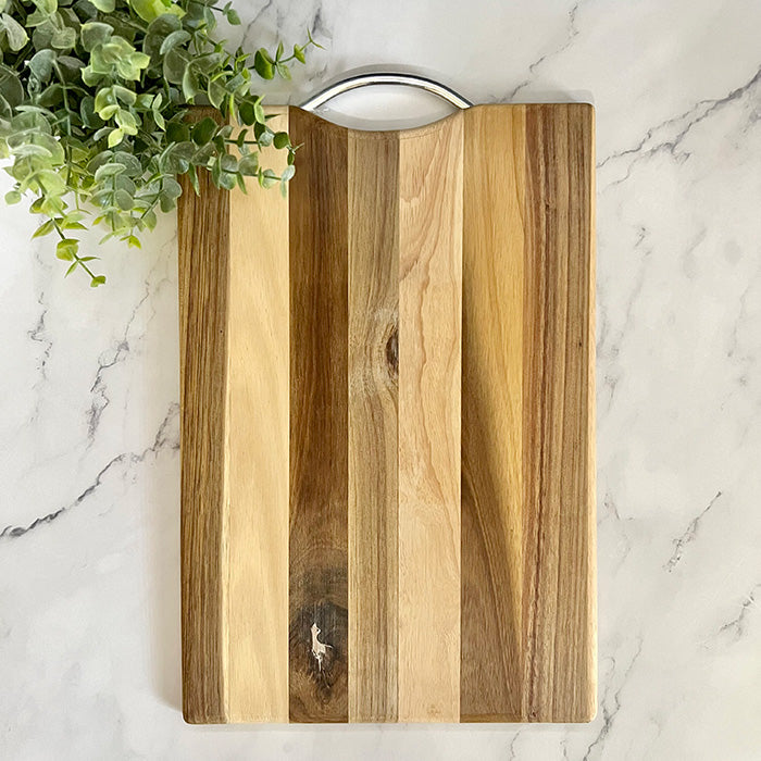 An acacia wood cutting board with a metal handle, displayed on a marble surface with a small green plant in the background.