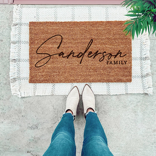 A personalized coir doormat with a name engraved on it, placed on a doorstep with a person wearing white shoes and blue jeans standing in front of it.