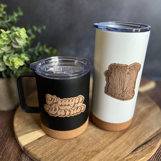 Two insulated drinkware cups, one tumbler and one travel mug, with wood veneer stickers, one black and one white, on a wooden board with a plant in the background.