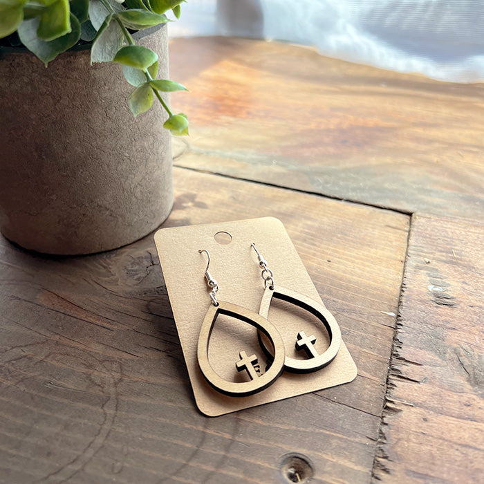 A pair of brown wooden teardrop cross earrings displayed on a card, placed on a wooden surface.