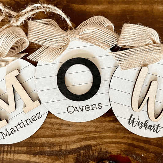 Hand-painted Christmas tree ornaments with Name text in black and natural wood on a white shiplap background, attached to a burlap bow.