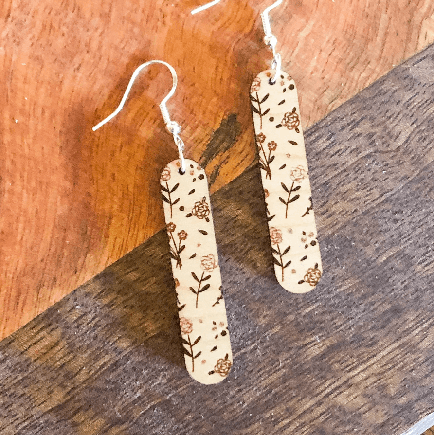 A pair of beige floral wooden dangle earrings with sterling silver hooks, displayed against a wooden background.