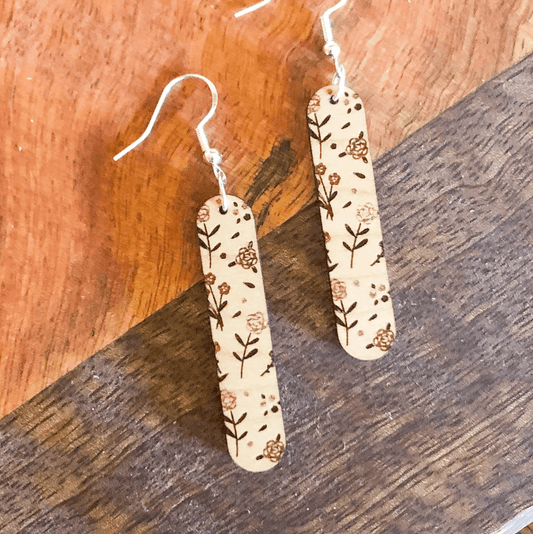 A pair of beige floral wooden dangle earrings with sterling silver hooks, displayed against a wooden background.