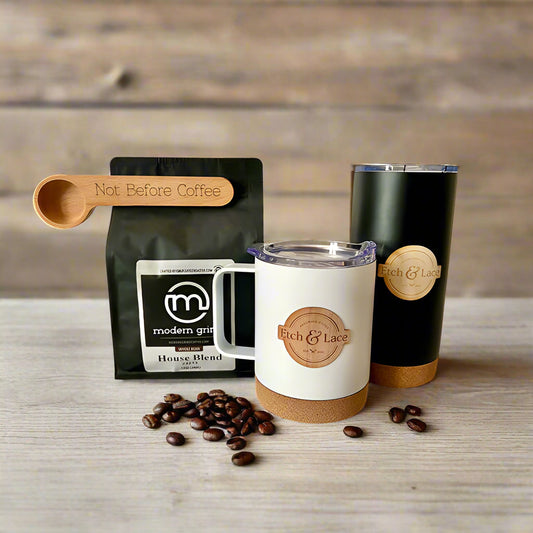 A gift set of coffee and drinkware, including a bag of Modern Grind coffee, an engraved coffee scoop, and a white travel mug with a wood veneer sticker, alongside coffee beans scattered on a wooden surface.