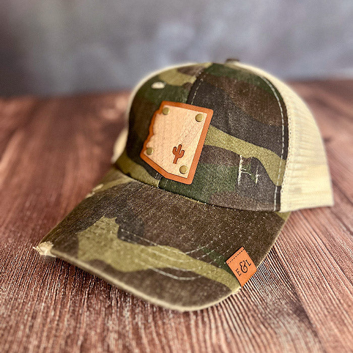 A camouflaged baseball cap with a leather patch featuring the Arizona state shape with a cutout of a cactus, designed for ponytail wear.