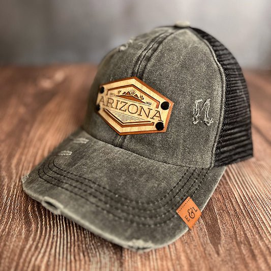 A black distressed ponytail hat with a 'Arizona' desert theme patch on wood veneer and vegan leather.