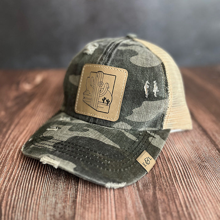 A grey camouflage ponytail hat with a laser-etched patch on the front featuring an AZ desert scene.