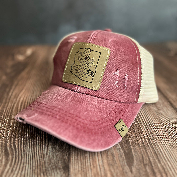 A red and beige ponytail hat with a laser-etched vegan leather patch featuring a cactus and desert scene, and an adjustable velcro closure.
