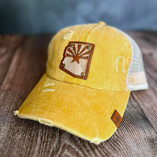A yellow distressed ponytail hat with a wood veneer patch shaped like the Arizona state and flag, and a leather patch on it.