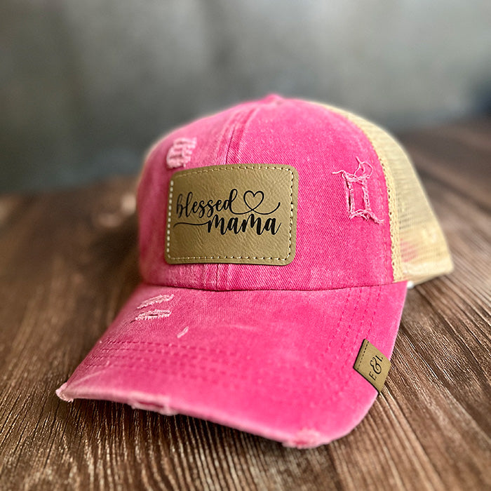 A pink trucker hat with a vegan leather patch that reads 'blessed mama', featuring a ponytail back and an adjustable velcro closure.