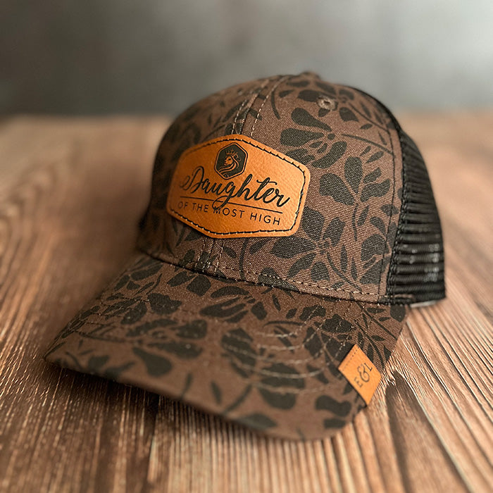 A brown ponytail hat with a floral pattern and a vegan leather patch that reads 'Daughter of the Most High' on the front.
