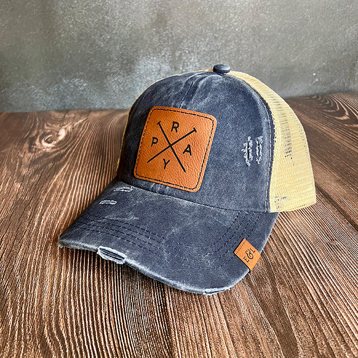 A navy and beige ponytail hat with a vegan leather patch that has 'PRAY' engraved on it.