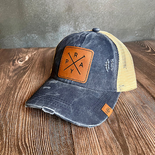 A navy and beige ponytail hat with a vegan leather patch that has 'PRAY' engraved on it.