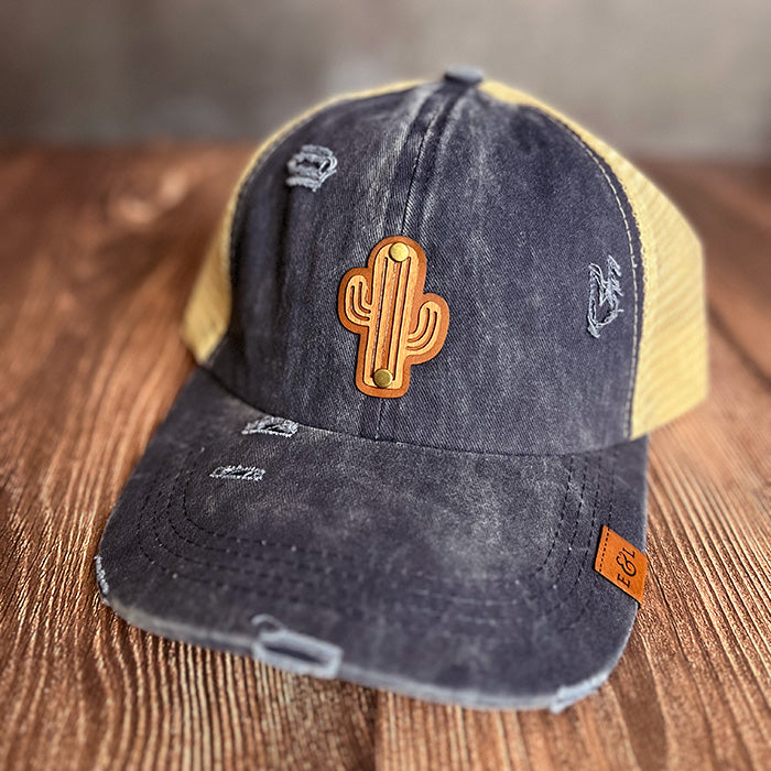 A distressed navy and beige ponytail hat with a wood veneer patch in the shape of a saguaro cactus.