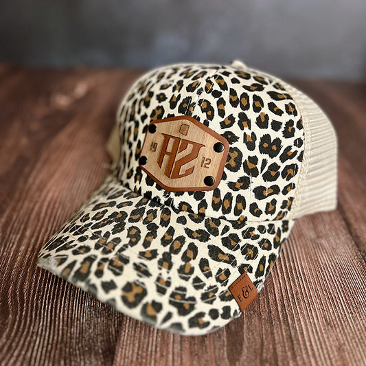 A beige and brown leopard print trucker hat with a wood veneer patch and a vegan leather patch featuring statehood details, displayed on a wooden surface.
