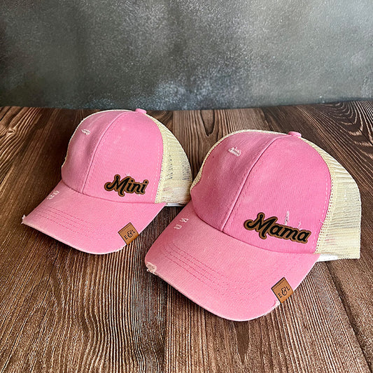 Two pink ponytail hats, one adult-sized and one child-sized, with 'Mama' and 'Mini' text engraved on a vegan leather patch.