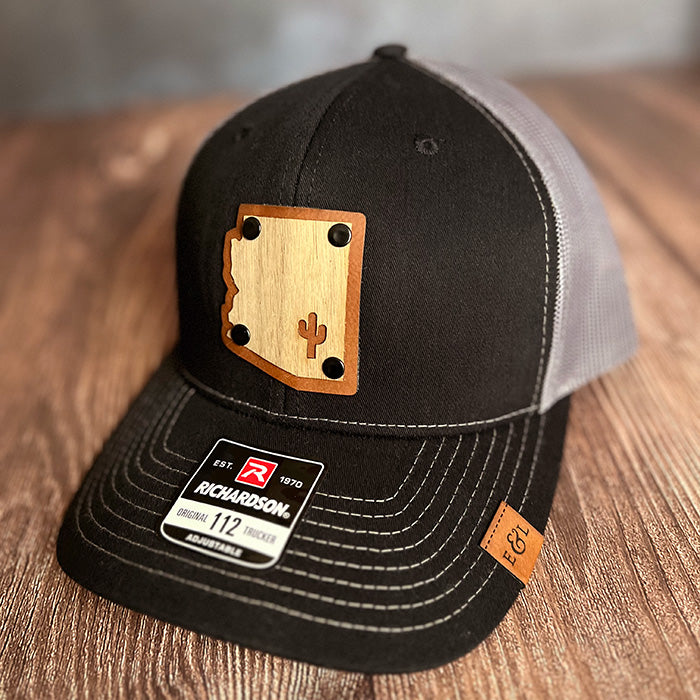 A black and gray trucker hat with a wood veneer patch featuring the Arizona State shape and a cactus design, finished with rivets.