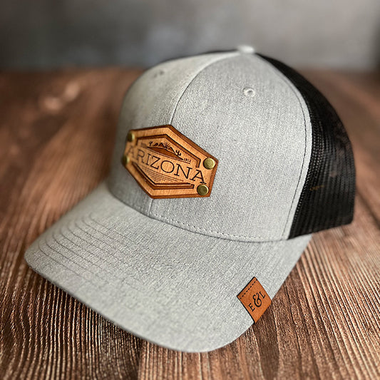 A gray and black trucker hat with a wood veneer and leather patch featuring an Arizona desert theme.
