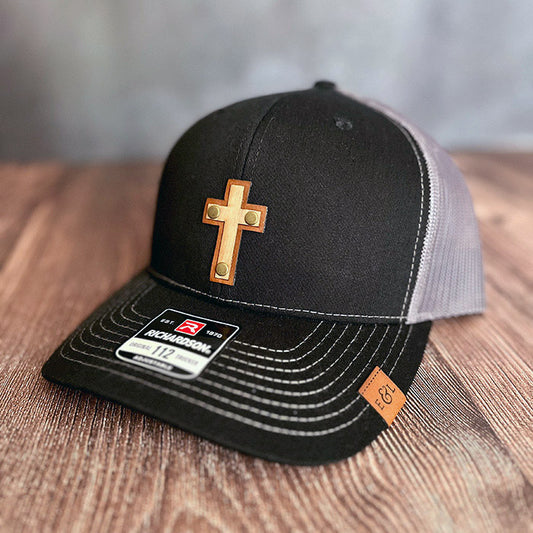 A black and gray Richardson 112 trucker hat with a cross-shaped wood veneer patch and a leather patch on the front.
