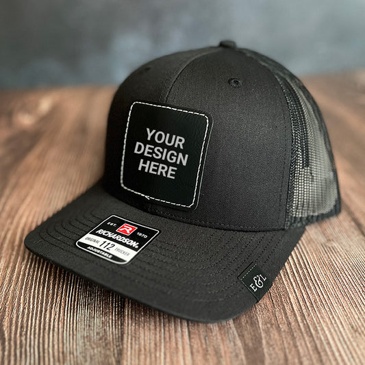 A black trucker hat with a custom patch design that reads 'YOUR DESIGN HERE' on the front, featuring a mesh back and an adjustable snapback closure. Richardson 112