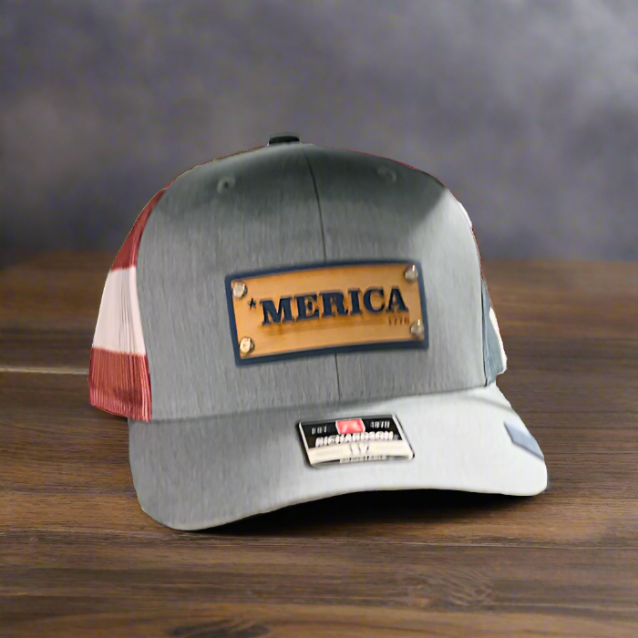 A gray stars and stripes Richardson 112 trucker hat with a wood veneer and leather riveted patch that has '*MERICA' laser-etched on it, displayed on a wooden surface.
