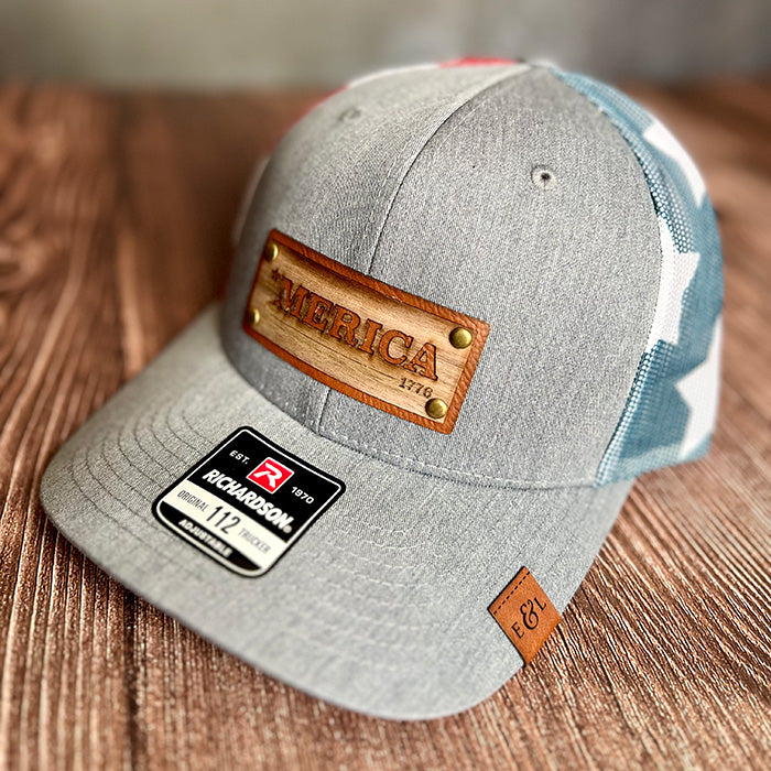 A gray stars and stripes Richardson 112 trucker hat with a wood veneer and leather riveted patch that has '*MERICA' laser-etched on it, displayed on a wooden surface.