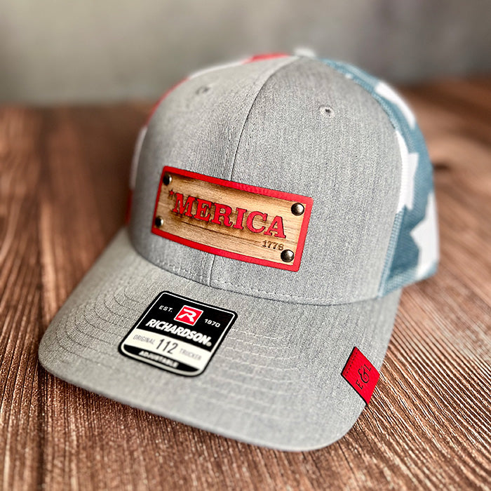 A gray stars and stripes Richardson 112 trucker hat with a wood veneer and leather riveted patch that has '*MERICA' laser-etched on it, displayed on a wooden surface.