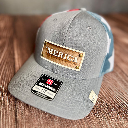 A gray stars and stripes Richardson 112 trucker hat with a wood veneer and leather riveted patch that has '*MERICA' laser-etched on it, displayed on a wooden surface.
