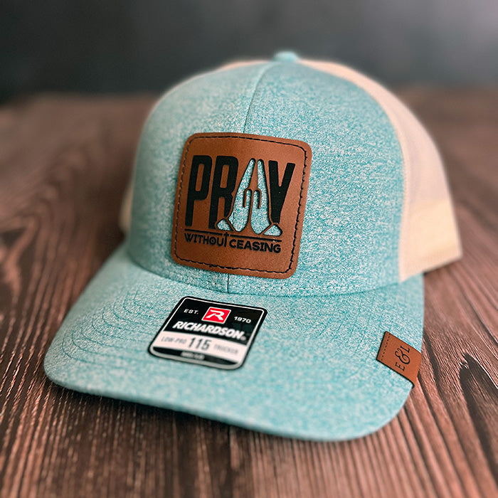 A teal and beige trucker hat with a vegan leather patch that has the text 'PRAY Without Ceasing' and a cutout of praying hands revealing the hat color underneath.
