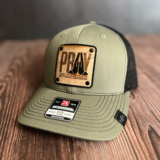 A green and black trucker hat with a vegan leather patch that has the text 'PRAY Without Ceasing' and a cutout of praying hands on veneer wood revealing the patch color underneath.