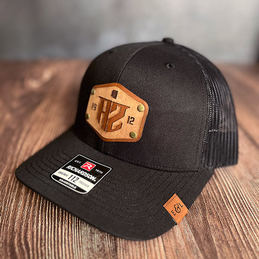 A black Richardson 112 trucker hat with a wood veneer and leather patch featuring state abbreviation and statehood date, with a visible label showing the product details.
