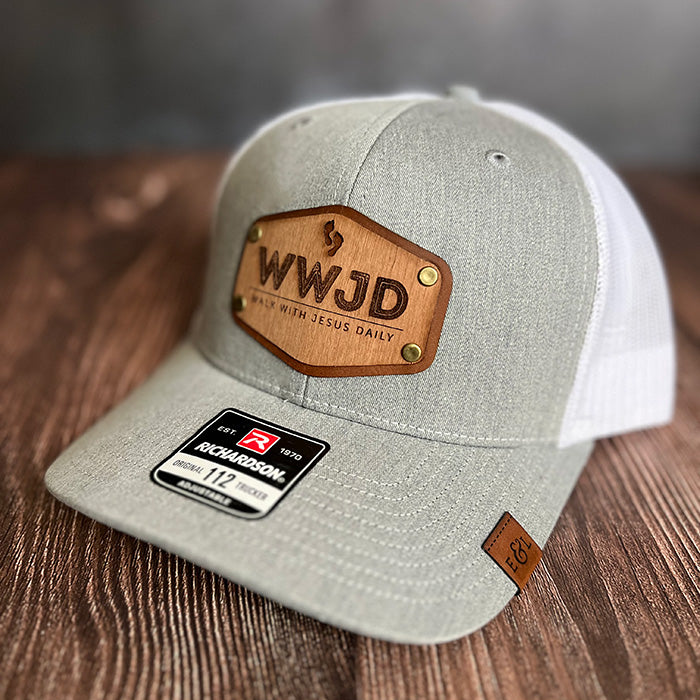 A gray and white trucker hat with a wood veneer and leather patch featuring the WWJD (Walk with Jesus Daily) message, and a label showing the brand and size.