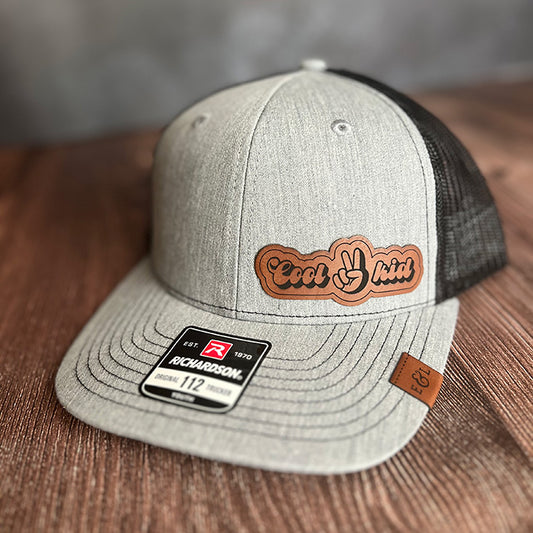Youth-size gray trucker cap with 'Cool Kid' text logo on the front and a leather strap, featuring an adjustable strap and a mesh back.