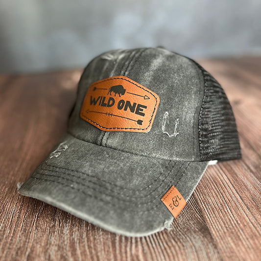 A vintage black youth trucker hat with a leather patch that reads 'WILD ONE' on the front. The hat features a mesh back and an adjustable strap.