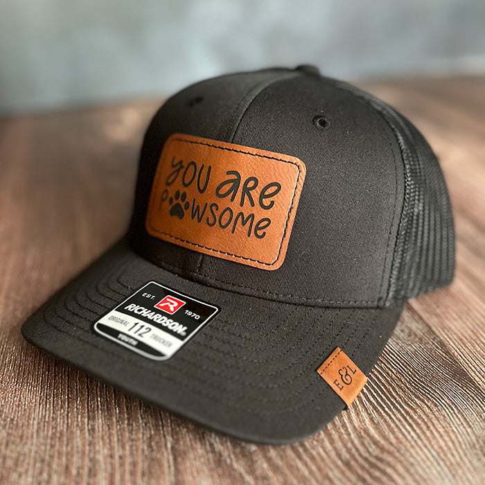 A youth-sized black trucker hat with a brown leather patch that has the phrase 'You are Pawsome' engraved on it, featuring a paw print design.