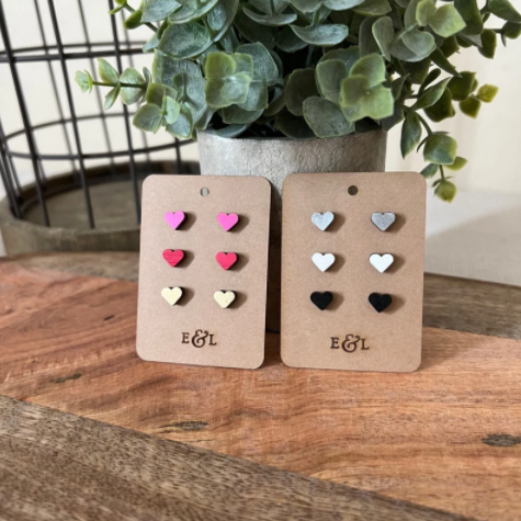 A pair of heart-shaped stud earrings in pink and white colors, displayed on a wooden surface with a plant in the background.