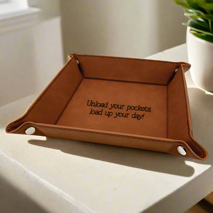 A brown leatherette key tray with silver snaps, placed on a white surface. The tray has a sleek design and includes a textual phrase that reads 'Unload your pockets and load up your day!'.