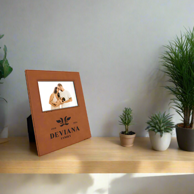 A brown leatherette picture frame with a photo of a couple displayed, placed on a wooden shelf with potted plants and decorative items around.