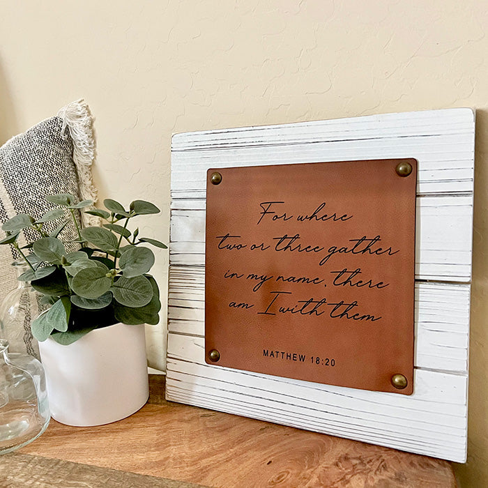 A rustic Scripture Wall Sign with white wood shiplap background and a brown vegan leather plaque featuring an engraved scripture passage. The sign is mounted on a wall on a shelf alongside a plant and a textured white object.