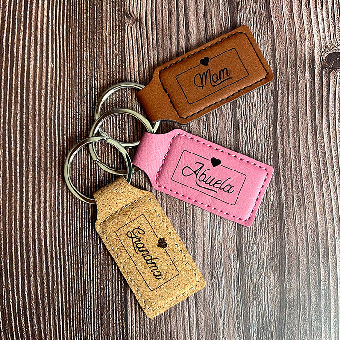 Three personalized vegan leather keychains in brown, pink, and cork, each with a different name engraved on them, displayed on a wooden surface.