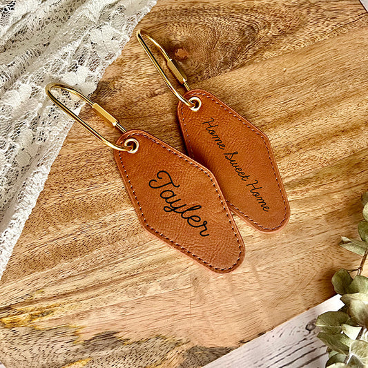 Two vintage motel vegan leather keychains with the word a name printed on them and home sweet home displayed on a wooden surface.