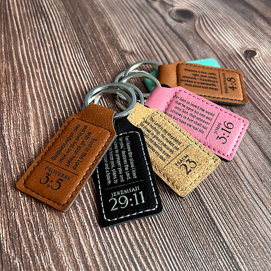 A collection of vegan leather keychains in black, brown, cork, pink, and teal colors, laser engraved with scripture verses.