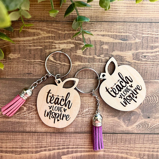 Two wooden apple-shaped keychains with the phrases 'Teach, Love, Inspire' engraved on them, one with a pink tassel and the other with a purple tassel, displayed on a wooden surface.