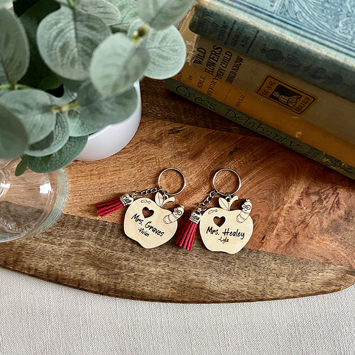 Two wooden apple-shaped keychains with personalized names and cutouts in the shape of worms, accompanied by colorful tassels, displayed on a wooden surface.