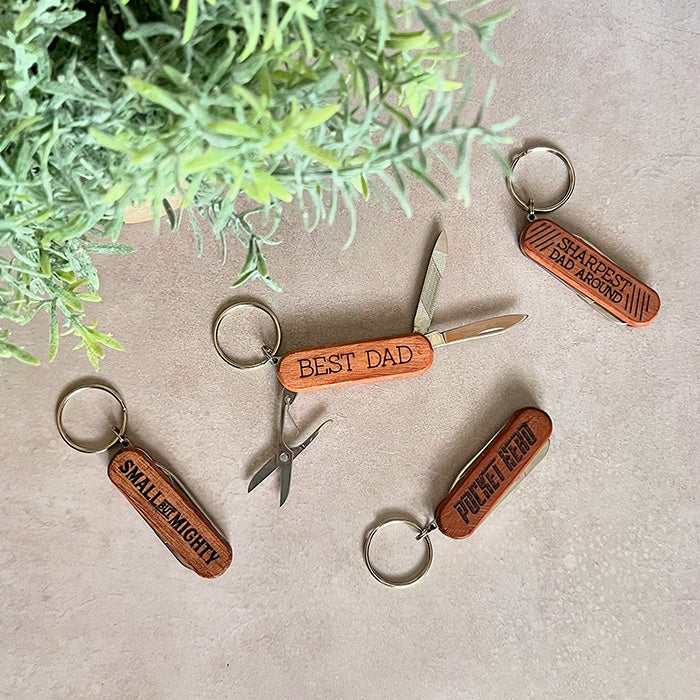 A collection of wooden pocket knives with keychain attachments. Each knife has a different inscription such as 'Best Dad' and 'Pocket Hero', and some are all partially open, showing the blade and the handle.