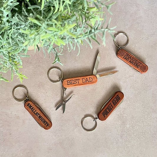 A collection of wooden pocket knives with keychain attachments. Each knife has a different inscription such as 'Best Dad' and 'Pocket Hero', and some are all partially open, showing the blade and the handle.
